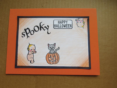 A halloween card matted on orange background with a the words spooky and happy halloween stamped on the front the card. A kewpie baby and a cat in a jack o lantern sits under the words